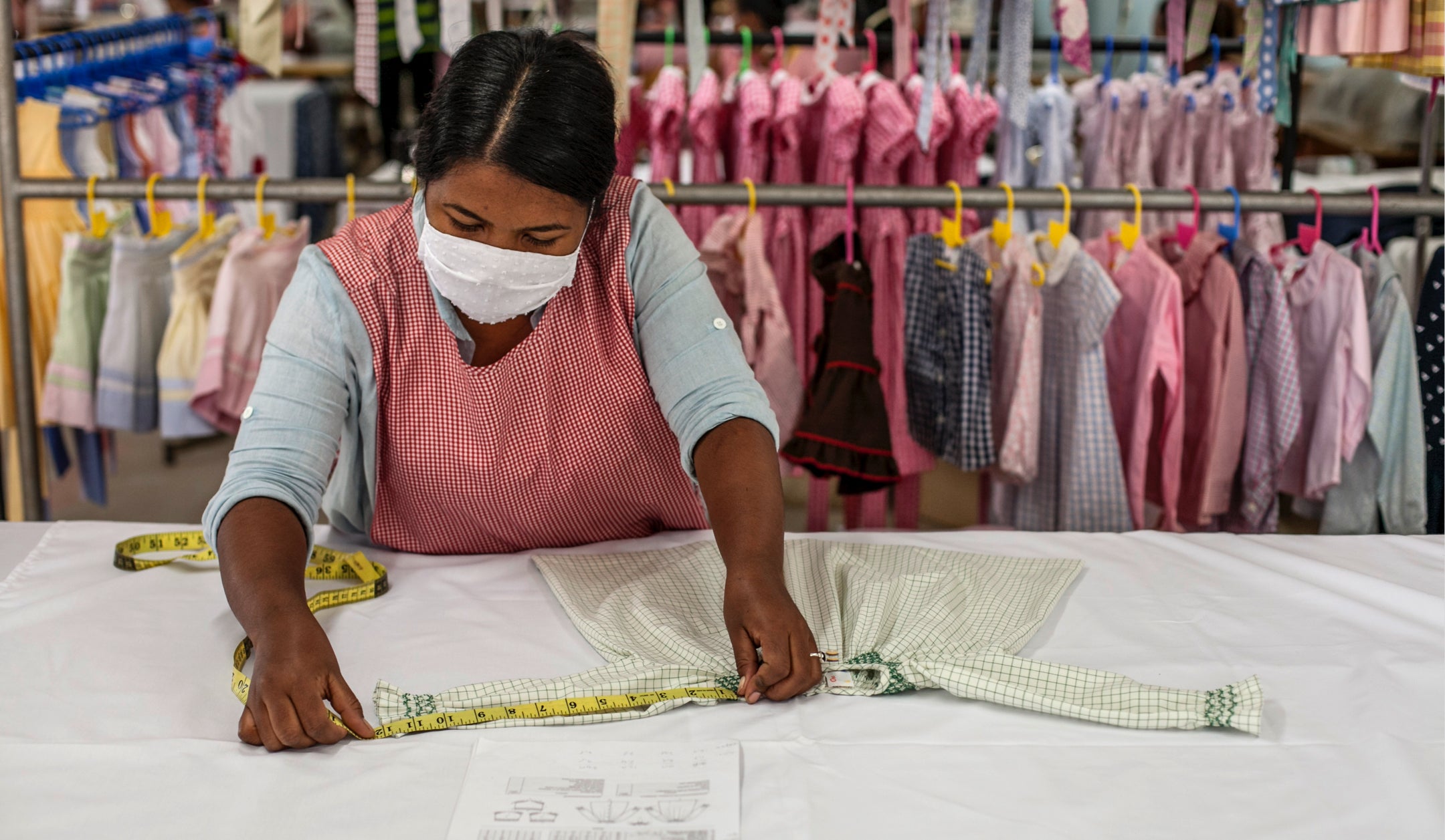 Women with mask measuring Willa Dress with measuring tape and clothes hanging in background.